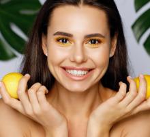Young woman with bright yellow eyeshadow, smiling and holding lemons near face.